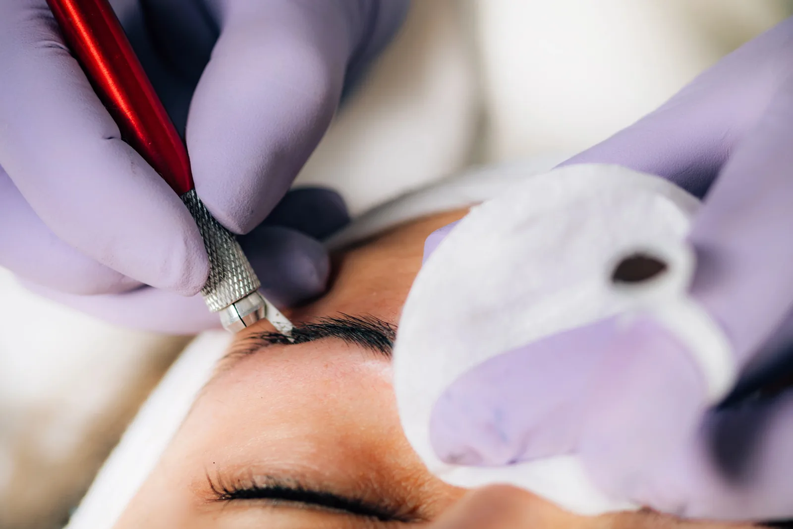 PMU-artiest met paarse handschoenen zet met een microbladingpen haarstrepen in een wenkbrauw.