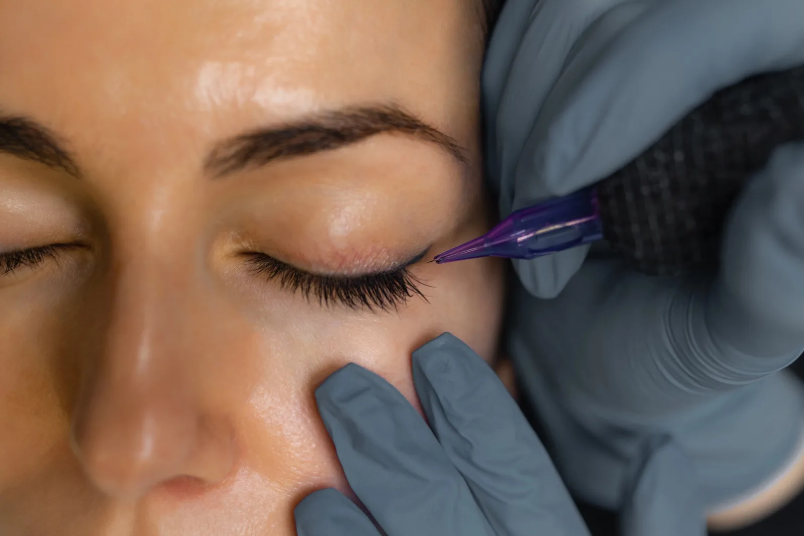 PMU-artiest zet met een fijne naald een wimperlijn tussen de wimpers van een klant met gesloten ogen.