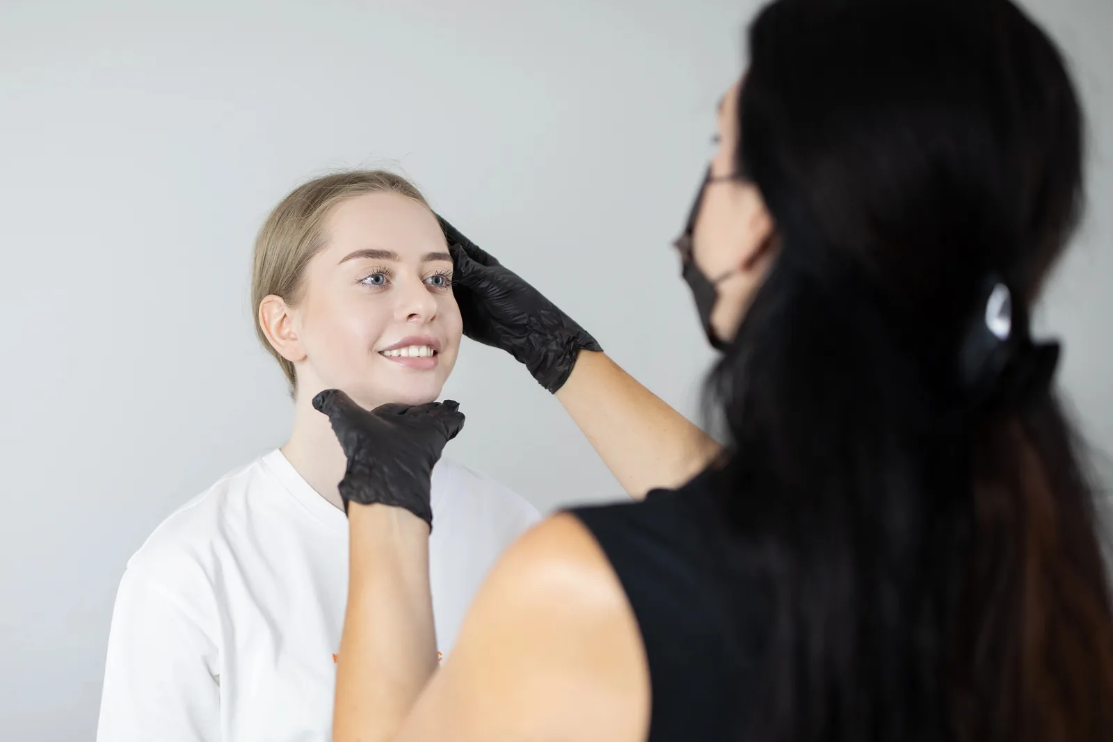Lachende klant en PMU-artiest met mondmasker bij een controle van de wenkbrauwen in de salon.