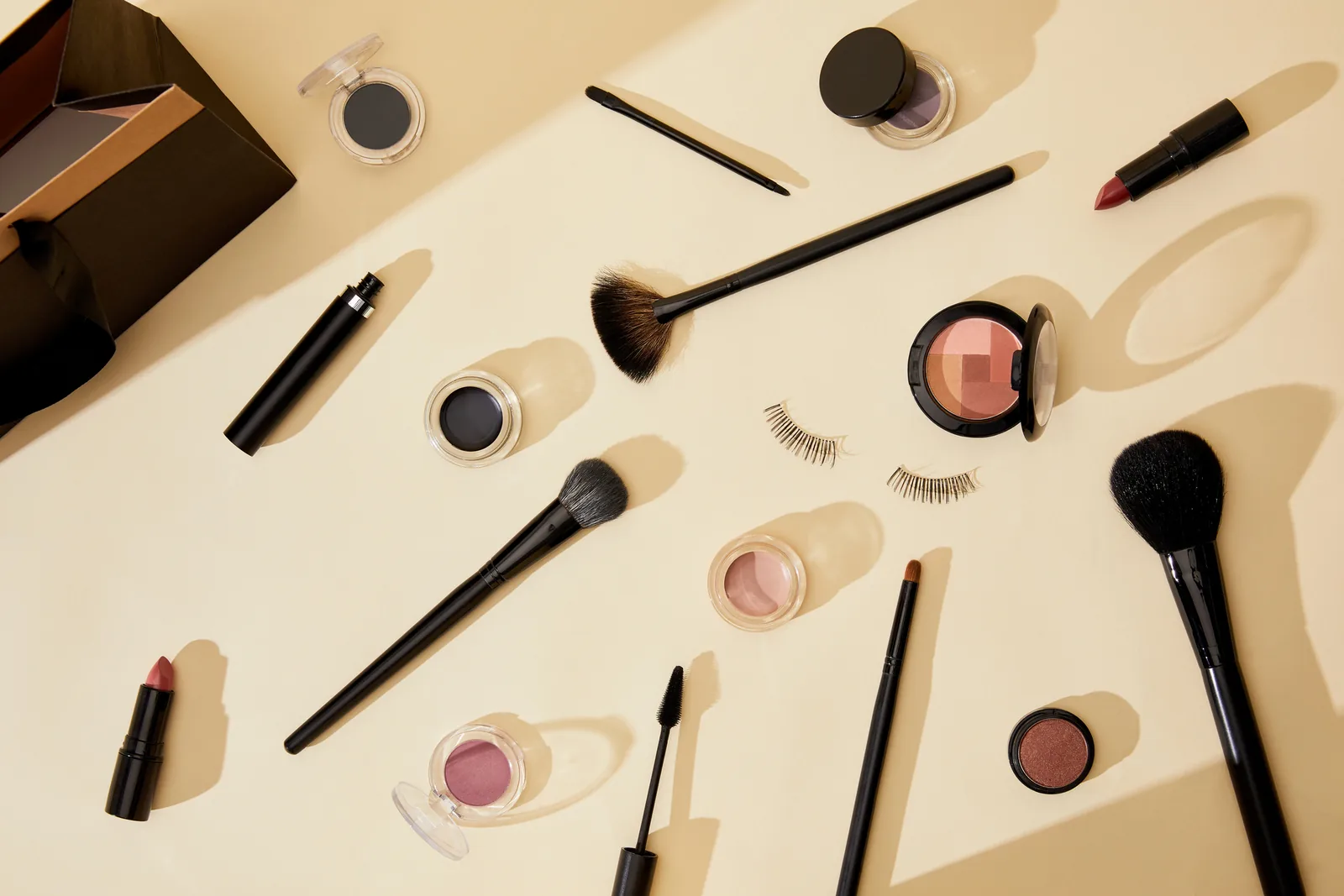 Bovenaanzicht van make-up, penselen, mascara en valse wimpers op een beige ondergrond.