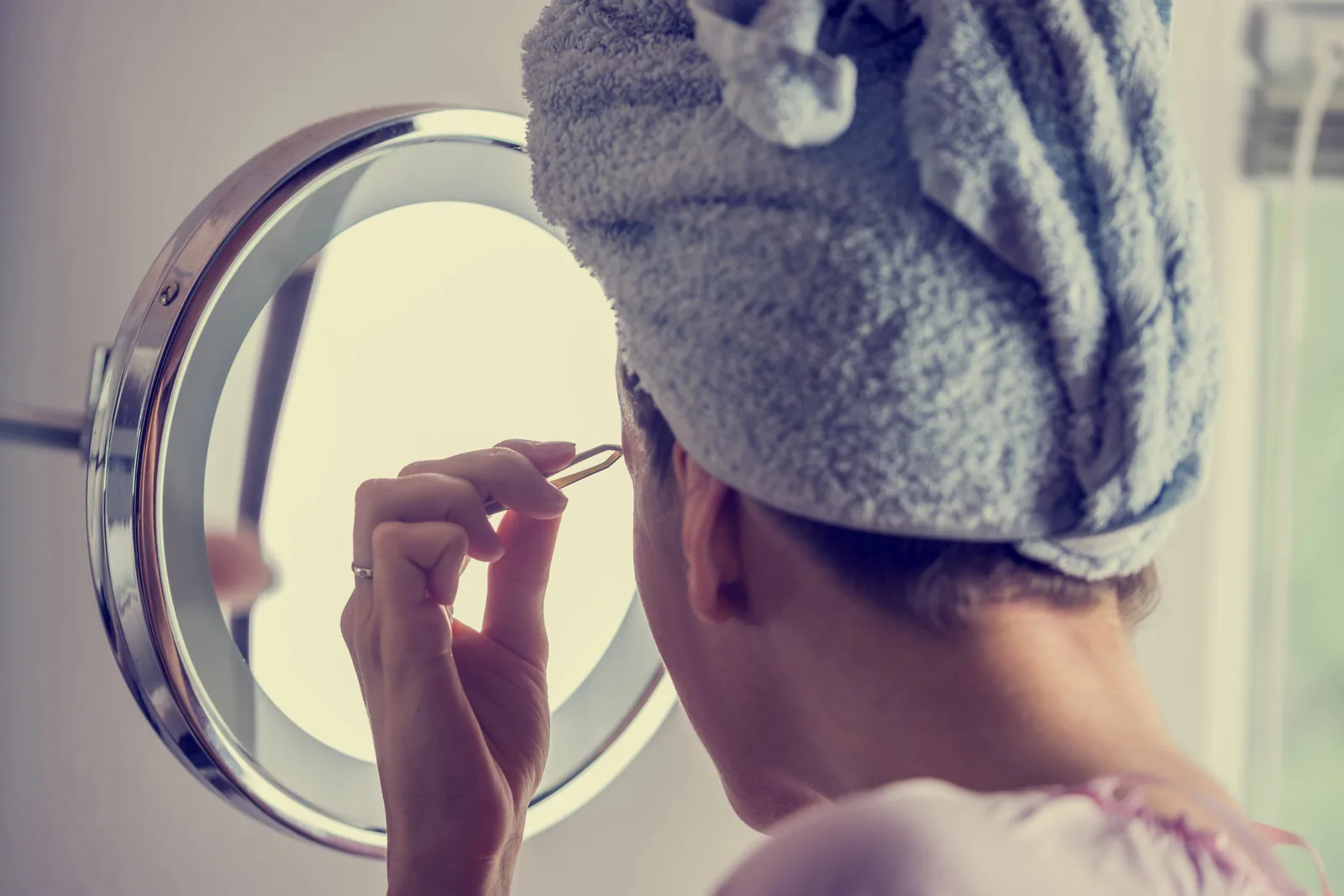 Vrouw met een handdoek rond het haar epileert haar wenkbrauwen voor de spiegel.